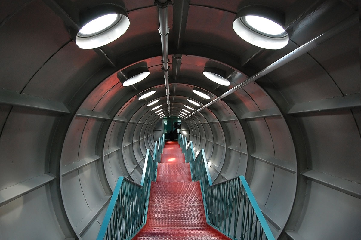 The Atomium tunnel with white circular lights in the roof, red steps descending down to an open door, with grey rounded walls and blue handrails